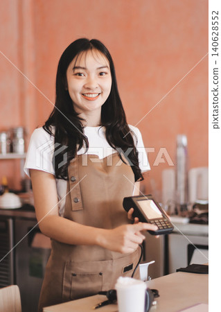 Friendly Asian female barista receives a credit card from a customer for payment. Both women are smiling in a warmly lit coffee shop with a rustic interior. Cashless payments. Small family business Friendly Asian female barista receives a credit card from a customer for payment. Both women are smiling in a warmly lit coffee shop with a rustic interior. Cashless payments. Small family business 140628552