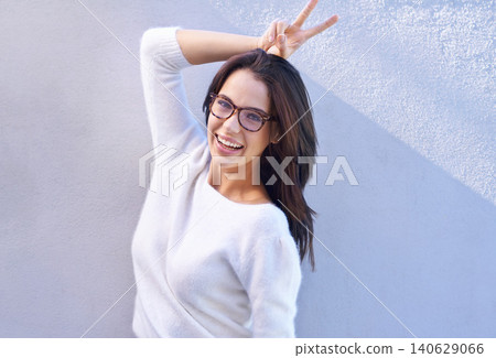 Portrait, fashion and happy with bunny ears woman on wall background for comedy, fun or humor. Smile, glasses and hand gesture with confident young person in trendy or casual clothing outfit Portrait, fashion and happy with bunny ears woman on wall background for comedy, fun or humor. Smile, glasses and hand gesture with confident young person in trendy or casual clothing outfit 140629066