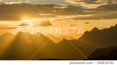Brilliant sun rays stream through clouds above the Dolomite mountains at sunset. Glowing alpine landscape with layered peaks, atmospheric haze and spectacular natural light. Brilliant sun rays stream through clouds above the Dolomite mountains at sunset. Glowing alpine landscape with layered peaks, atmospheric haze and spectacular natural light. 140630782
