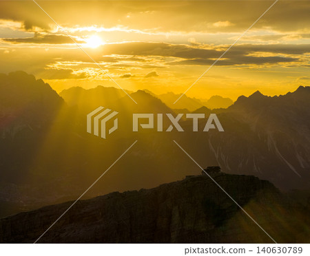 Brilliant sun rays stream through clouds above the Dolomite mountains at sunset. Glowing alpine landscape with layered peaks, atmospheric haze and spectacular natural light. 140630789