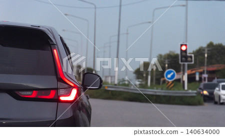 Rear corner perspective of a modern black car waiting on a asphalt roadway. Traffic signals and street lamps line the highway divider section further ahead. Rear corner perspective of a modern black car waiting on a asphalt roadway. Traffic signals and street lamps line the highway divider section further ahead. 140634000