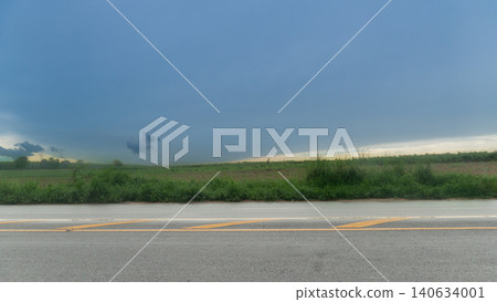 Horizontal or Landscape showing an empty wet rainy asphalt roadway. Beneath a wide dark storm cloud formation. Lush green grassy fields border the modern concrete landscape layout. Horizontal or Landscape showing an empty wet rainy asphalt roadway. Beneath a wide dark storm cloud formation. Lush green grassy fields border the modern concrete landscape layout. 140634001