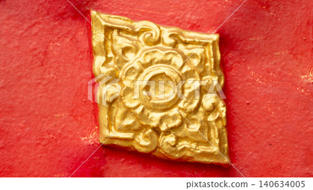 Close-up view showing a single square golden flower-shaped Lai Kanok. Thai ornament ornament attached firmly to a rough-textured red stucco wall panel background. Close-up view showing a single square golden flower-shaped Lai Kanok. Thai ornament ornament attached firmly to a rough-textured red stucco wall panel background. 140634005
