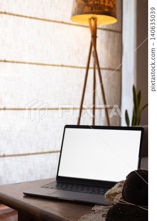 Laptop with blank screen on wooden table beside yarn, knitting needles and a partially finished crochet piece, cozy home workspace suggesting online learning and creative hobby Laptop with blank screen on wooden table beside yarn, knitting needles and a partially finished crochet piece, cozy home workspace suggesting online learning and creative hobby 140635039