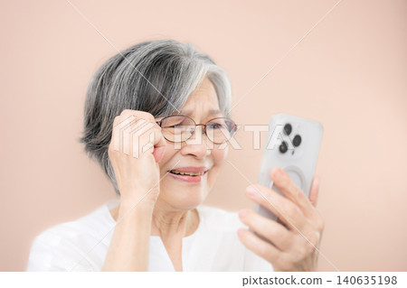 A gray-haired senior woman wearing reading glasses and looking at her smartphone against a pink background; the text is difficult to read, suggesting she might be a scammer. A gray-haired senior woman wearing reading glasses and looking at her smartphone against a pink background; the text is difficult to read, suggesting she might be a scammer. 140635198