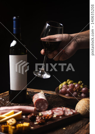 Vertical shot of a hand holding a glass of red wine while enjoying a charcuterie board on a rustic wooden table, celebrating a luxurious lifestyle with a delicious appetizer. 140635208
