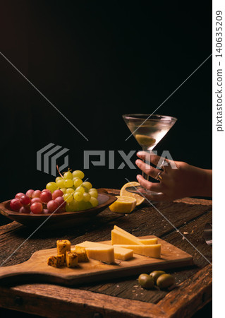 A dry gin martini, made with premium spirits, is held by a woman's hand, with fresh grapes, hints of lemon, and a gourmet cheese board creating a refined gourmet experience. A dry gin martini, made with premium spirits, is held by a woman's hand, with fresh grapes, hints of lemon, and a gourmet cheese board creating a refined gourmet experience. 140635209