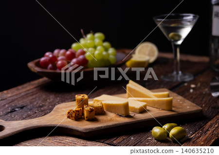 Still life displaying gourmet appetizers including cheeses, grapes, olives, and a drink with lemon on a rustic wooden table 140635210