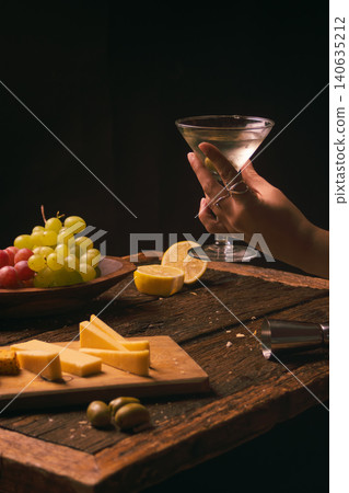 Hand holding classic martini with olives beside a wooden bowl of green and red grapes, sliced cheese and lemon on a rustic table, suggesting a relaxed elegant gathering Hand holding classic martini with olives beside a wooden bowl of green and red grapes, sliced cheese and lemon on a rustic table, suggesting a relaxed elegant gathering 140635212