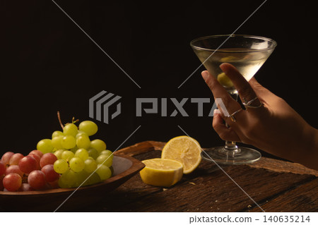 A person holds a chilled martini with an olive next to a cheese and charcuterie board, enjoying a luxury cocktail and gourmet appetizers. A person holds a chilled martini with an olive next to a cheese and charcuterie board, enjoying a luxury cocktail and gourmet appetizers. 140635214