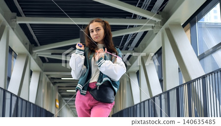 Caucasian woman posing for photo on bridge styling varsity jacket and backpack, hand resting on railing, expressive gaze toward camera, geometric metal beams and glass background, fashion content tone Caucasian woman posing for photo on bridge styling varsity jacket and backpack, hand resting on railing, expressive gaze toward camera, geometric metal beams and glass background, fashion content tone 140635485