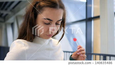 Close portrait of young woman near window holding lollipop with thoughtful expression, soft daylight illuminating face and textured sweater, intimate quiet mood in transit corridor. Close portrait of young woman near window holding lollipop with thoughtful expression, soft daylight illuminating face and textured sweater, intimate quiet mood in transit corridor. 140635486