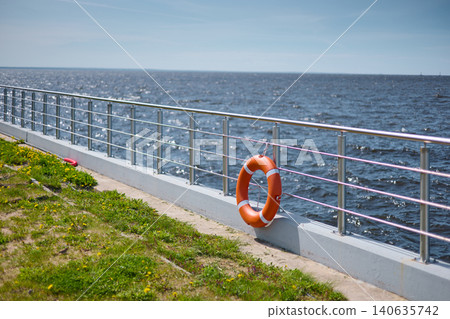 An orange-colored lifebuoy hangs on the embankment fence in sunny weather, finland gulf, with the sea in the background. 140635742