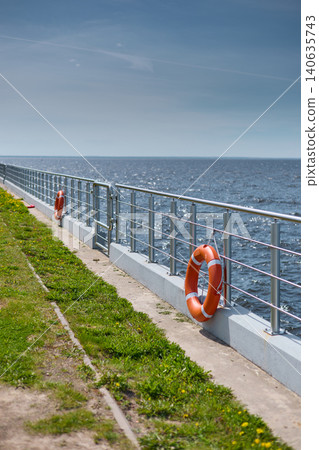 An orange-colored lifebuoy hangs on the embankment fence in sunny weather, finland gulf, with the sea in the background. 140635743
