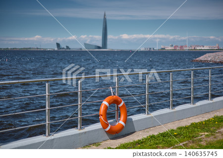 An orange-colored lifebuoy hangs on the embankment fence in sunny weather, finland gulf, with the sea and a skyscraper in the background. An orange-colored lifebuoy hangs on the embankment fence in sunny weather, finland gulf, with the sea and a skyscraper in the background. 140635745