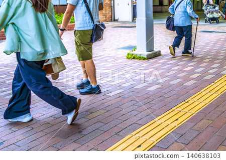 In the urban landscape of Yokohama, Japan, an elderly woman with a cane (wearing a mask) scatters feed for pigeons... lives intersect. A double stroller... young people... 140638103