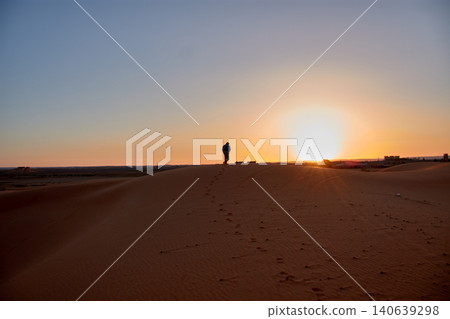 Silhouette of a tourist exploring the golden dunes under the warm light of the setting sun in Merzouga, Morocco Silhouette of a tourist exploring the golden dunes under the warm light of the setting sun in Merzouga, Morocco 140639298