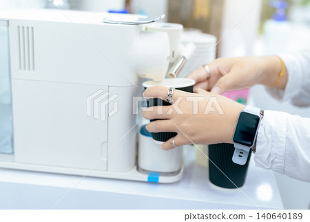Woman making a cup of hot coffee with capsule coffee machine. Woman holding black cup taking coffee from a capsule Modern machine on the table. Espresso maker. Morning drink. International coffee day. Woman making a cup of hot coffee with capsule coffee machine. Woman holding black cup taking coffee from a capsule Modern machine on the table. Espresso maker. Morning drink. International coffee day. 140640189