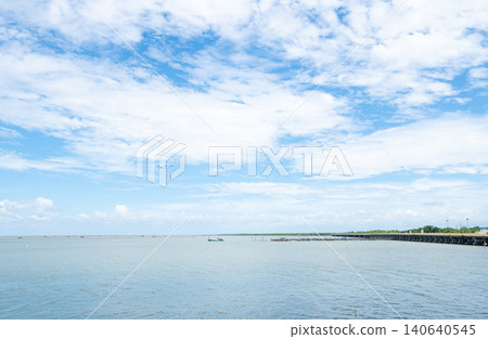 Wide coastal seascape with calm ocean water and dramatic cloud filled blue sky, peaceful tropical shoreline landscape. Distant pier and open horizon during summer day. Coastal travel destination. 140640545