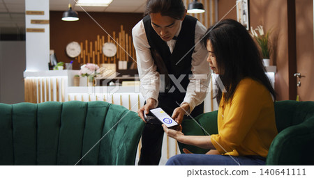 Tourist using NFC payment to buy coffee cup from hotel bar, employee bringing drink in lounge area, Asian woman relaxing in lobby, making mobile phone transaction at POS terminal, handheld shot. Tourist using NFC payment to buy coffee cup from hotel bar, employee bringing drink in lounge area, Asian woman relaxing in lobby, making mobile phone transaction at POS terminal, handheld shot. 140641111
