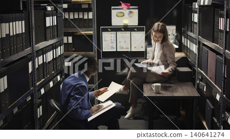 In room crammed with clues and records, Caucasian female investigator sits on table analyzing evidence with police officer, African American inspector hands over case data to policewoman with laptop. In room crammed with clues and records, Caucasian female investigator sits on table analyzing evidence with police officer, African American inspector hands over case data to policewoman with laptop. 140641274