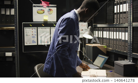 Devoted private investigator pulls files from shelf to conduct a comprehensive investigation, with laptop and research paperwork, African American agent analyzes evidence and gathers information. Devoted private investigator pulls files from shelf to conduct a comprehensive investigation, with laptop and research paperwork, African American agent analyzes evidence and gathers information. 140641278