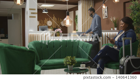 Tourist asks for help and room card in lobby, ringing bell to call for receptionist at front desk and give assistance, African American person with trolley bags requesting access for room. 140641360