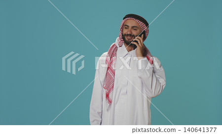 Islamic guy in robe texting on phone while posing with confidence over blue background. Islamic guy in robe texting on phone while posing with confidence over blue background. 140641377