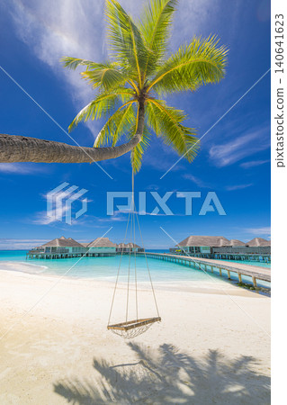 Relaxing beach swing hanging from a palm tree above turquoise water with overwater bungalows beyond. Romantic Maldives paradise scene. Stunning summer travel background with white sand and blue sky Relaxing beach swing hanging from a palm tree above turquoise water with overwater bungalows beyond. Romantic Maldives paradise scene. Stunning summer travel background with white sand and blue sky 140641623