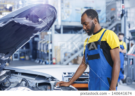 BIPOC specialist in car service using professional mechanical tool to repair broken motor, with trained specialist in garage fixing client automobile, ensuring optimal automotive performance. BIPOC specialist in car service using professional mechanical tool to repair broken motor, with trained specialist in garage fixing client automobile, ensuring optimal automotive performance. 140641674