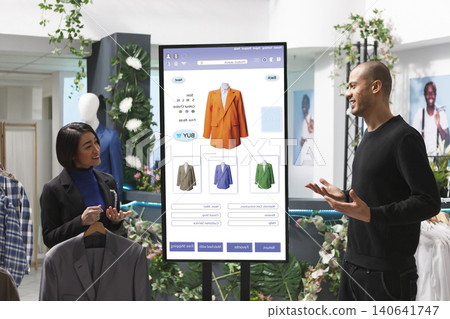 Store associate displays items to man in front of digital kiosk, selecting merchandise from clothing boutique, with male customer converses with female employee about products. Store associate displays items to man in front of digital kiosk, selecting merchandise from clothing boutique, with male customer converses with female employee about products. 140641747
