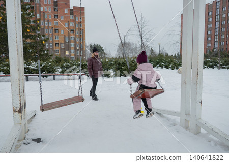 Child swinging toward camera in snow mid-air motion with visible swing chains, caregiver walking nearby in puffer coat, footprints on compacted pathway, overcast sky and apartment buildings Child swinging toward camera in snow mid-air motion with visible swing chains, caregiver walking nearby in puffer coat, footprints on compacted pathway, overcast sky and apartment buildings 140641822