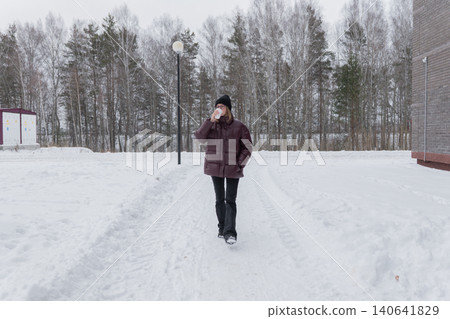 caucasian woman walking on snow path, maroon puffer and black beanie, holding cup, steady stride, pine tree background, cool overcast atmosphere, purposeful commute, fresh winter energy caucasian woman walking on snow path, maroon puffer and black beanie, holding cup, steady stride, pine tree background, cool overcast atmosphere, purposeful commute, fresh winter energy 140641829