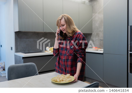 Blonde white woman talking on phone near dining table with fresh pie, multitasking between call and food prep. Plaid shirt and modern cabinetry frame a cozy but efficient kitchen scene. Expression Blonde white woman talking on phone near dining table with fresh pie, multitasking between call and food prep. Plaid shirt and modern cabinetry frame a cozy but efficient kitchen scene. Expression 140641834
