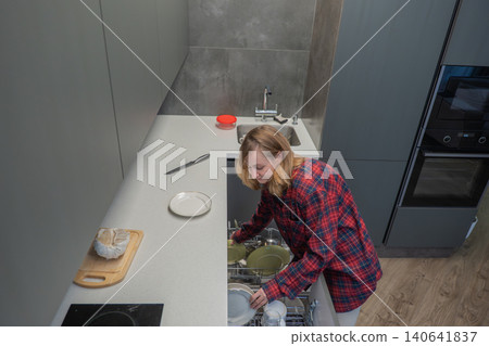 Young white woman unloading dishwasher racks in sleek kitchen after meal, placing plates into drying area while wearing cozy plaid shirt, warm indoor light, cookware and utensils nearby, relaxed Young white woman unloading dishwasher racks in sleek kitchen after meal, placing plates into drying area while wearing cozy plaid shirt, warm indoor light, cookware and utensils nearby, relaxed 140641837
