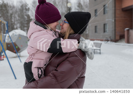 white aunt kissing daughters nose under snowy sky, tender forehead and cheek contact, warm puffer coats and mittens, playful grip on climbing ropes, playground structure and building behind, white aunt kissing daughters nose under snowy sky, tender forehead and cheek contact, warm puffer coats and mittens, playful grip on climbing ropes, playground structure and building behind, 140641850