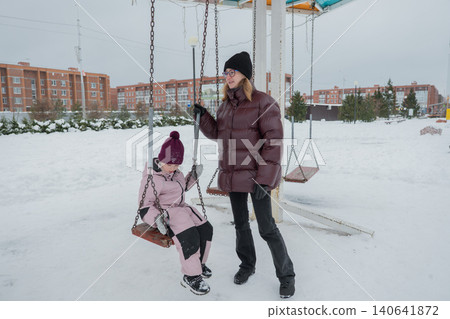 Smiling caucasian mother and daughter swinging together in snowy playground, mother in maroon puffer, child in pink coat and maroon hat, laughter and eye contact, suburban apartment skyline, family Smiling caucasian mother and daughter swinging together in snowy playground, mother in maroon puffer, child in pink coat and maroon hat, laughter and eye contact, suburban apartment skyline, family 140641872