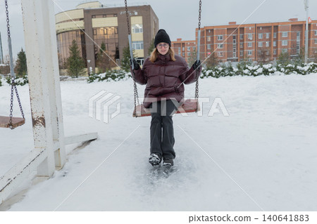Young caucasian woman sitting on swing in snowy park wearing maroon puffer and black beanie, relaxed smile, urban apartment blocks in background, chains creaking, footprints in fresh snow, leisure 140641883
