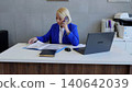 Businesswoman wearing a blue suit sitting at her desk, talking on the phone and checking documents, with a laptop and printer in the background 140642039