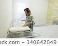 Woman airing pillow and duvet, home organizer arranging linen on drying rack near window, natural light fills room, soft textured wallpaper, tidy neutral decor, calm morning routine, sustainable 140642049