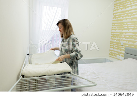 Woman airing pillow and duvet, home organizer arranging linen on drying rack near window, natural light fills room, soft textured wallpaper, tidy neutral decor, calm morning routine, sustainable Woman airing pillow and duvet, home organizer arranging linen on drying rack near window, natural light fills room, soft textured wallpaper, tidy neutral decor, calm morning routine, sustainable 140642049