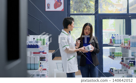 Shopper in pharmacy needing prescription drug to calm nausea feeling, asking pharmacist to help her pick best medicament, with knowledgeable pharmacologist assisting customer with product counsel. Shopper in pharmacy needing prescription drug to calm nausea feeling, asking pharmacist to help her pick best medicament, with knowledgeable pharmacologist assisting customer with product counsel. 140642052