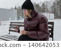 White woman reading messages on phone while holding coffee on snowy bench, maroon puffer, black beanie, glasses, pine forest background, quiet winter atmosphere, checking schedule, outdoor study 140642053