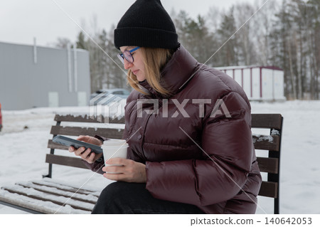 White woman reading messages on phone while holding coffee on snowy bench, maroon puffer, black beanie, glasses, pine forest background, quiet winter atmosphere, checking schedule, outdoor study White woman reading messages on phone while holding coffee on snowy bench, maroon puffer, black beanie, glasses, pine forest background, quiet winter atmosphere, checking schedule, outdoor study 140642053