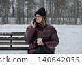 smiling woman on phone holding coffee, warm expression during friendly conversation, closeup portrait with glasses and knit beanie, maroon jacket contrasts snowy background, candid joyful moment 140642055