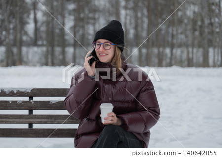smiling woman on phone holding coffee, warm expression during friendly conversation, closeup portrait with glasses and knit beanie, maroon jacket contrasts snowy background, candid joyful moment smiling woman on phone holding coffee, warm expression during friendly conversation, closeup portrait with glasses and knit beanie, maroon jacket contrasts snowy background, candid joyful moment 140642055