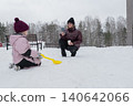 A white mother kneels with her daughter, snow shows the mother teaching her daughter shoveling techniques with a bright yellow shovel, folded coats and knitted hats, a snow-covered playground 140642066