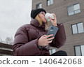 White woman sipping coffee, checking phone on park bench in snowy urban park, wearing burgundy puffer jacket and black beanie, relaxed smile, winter morning, hands wrapped around cup, social scroll, 140642068