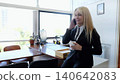 Blonde businesswoman in a suit smiling while talking on the phone and holding a coffee cup in her office 140642083