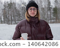 Woman in puffer jacket holding coffee, park setting with snowcovered trees, calm expression with round glasses and knit beanie, warm breath in crisp air, hands wrapped around cup, serene winter pause 140642086